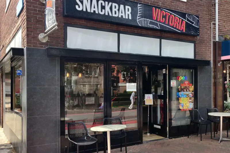 De gevel van Snackbar Victoria met het zwarte uithangbord en het terras voor de deur